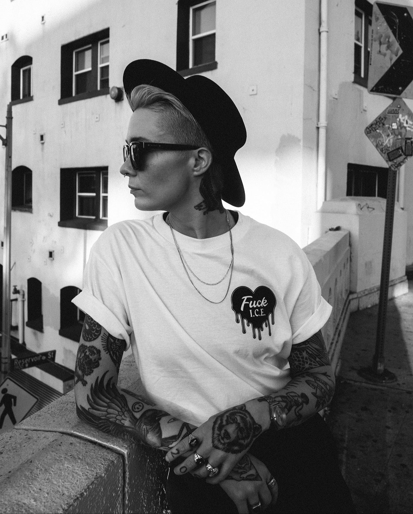 Person with tattoos wearing a white t-shirt with a graphic design, sitting on a bench in an urban setting.